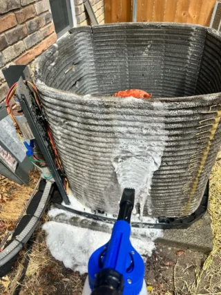 Technician using a blue pressure washer to clean a heavily soiled AC condenser coil caked with dirt and debris during maintenance in Mesquite, TX.