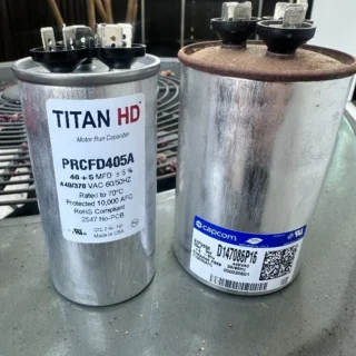 Two failed dual run capacitors side by side, showing TITAN HD and Capcom branded units with visible terminals and specifications labels.