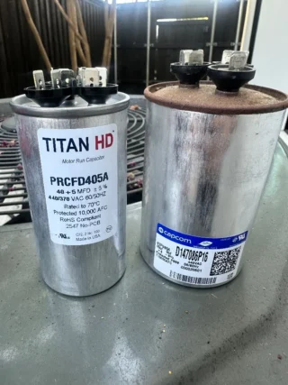 Two failed dual run capacitors side by side, showing TITAN HD and Capcom branded units with visible terminals and specifications labels.