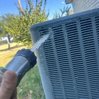 Technician spraying water on a clogged AC condenser coil during maintenance in Mesquite, TX