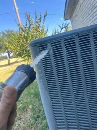 Technician spraying water on a clogged AC condenser coil during maintenance in Mesquite, TX