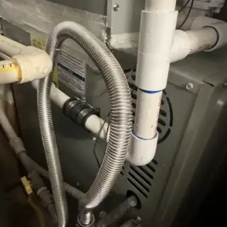 Close-up of an AC condensate drain line with metal flexible tubing and white PVC pipes showing water drainage system components.