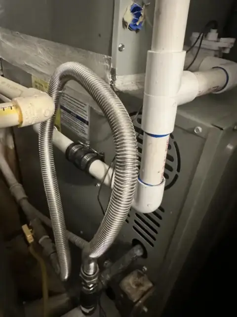 Close-up of an AC condensate drain line with metal flexible tubing and white PVC pipes showing water drainage system components.